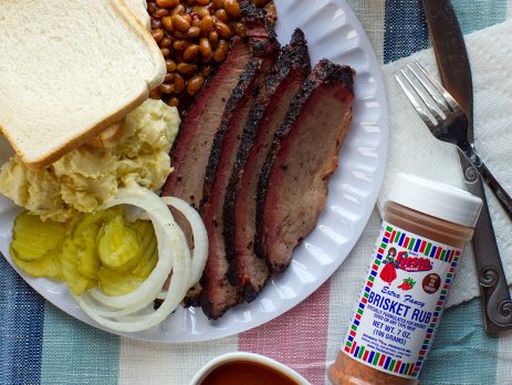 Plate of sliced brisket with beans, potato salad, pickles, onions, bread, and jar of Fiesta Brisket Rub.