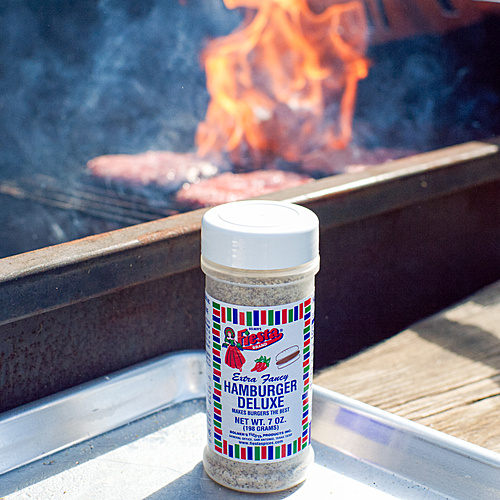 Close-up shot of medium size jar of Hamburger Deluxe next to a flaming backyard BBQ grill.