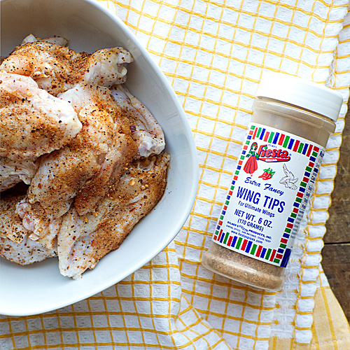 Seasoning the chicken wings using Fiesta Brand Wing Tips seasoning. Coating wings evenly and tossing them in a bowl before placing over hot coals on a backyard BBQ pit.