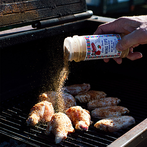 Fiesta Brand Wing Tips seasoning being sprinkled over raw chicken wings on a charcoal grill.